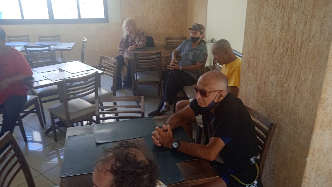 Na imagem, um grupo de idosos sentados em mesas e cadeiras, assistem a oficina. Um senhor na mesa da frente veste preto e usa óculos escuros, atrás dele um senhor de amarelo. Na mesa mais ao fundo, um senhor de máscara e boné, veste uma camisa azul e calças azuis. Ao lado dele, uma senhora veste uma camisa florida.